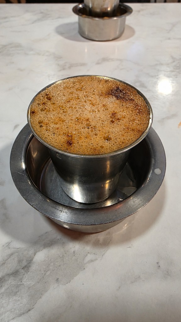Frothy coffee in a metal tumbler and saucer on a marble café table