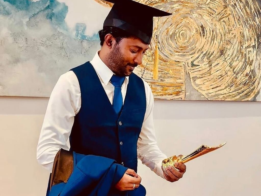 Thana wearing a graduation cap and blue waistcoat, looking down at a document in his hand.