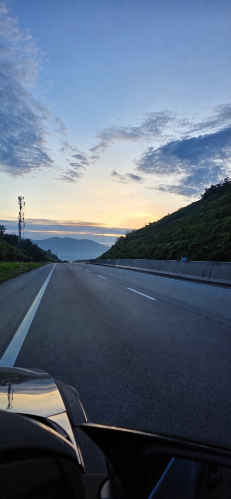 A highway between green hills beneath a brightening morning sky, seen from a car
