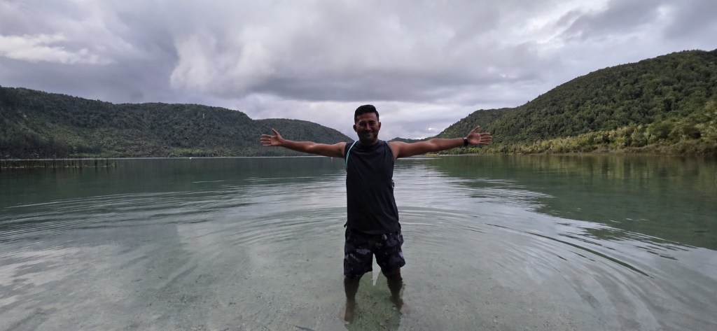 Thana standing with arms open in the water at Rotorua, New Zealand, with wooded hills behind him