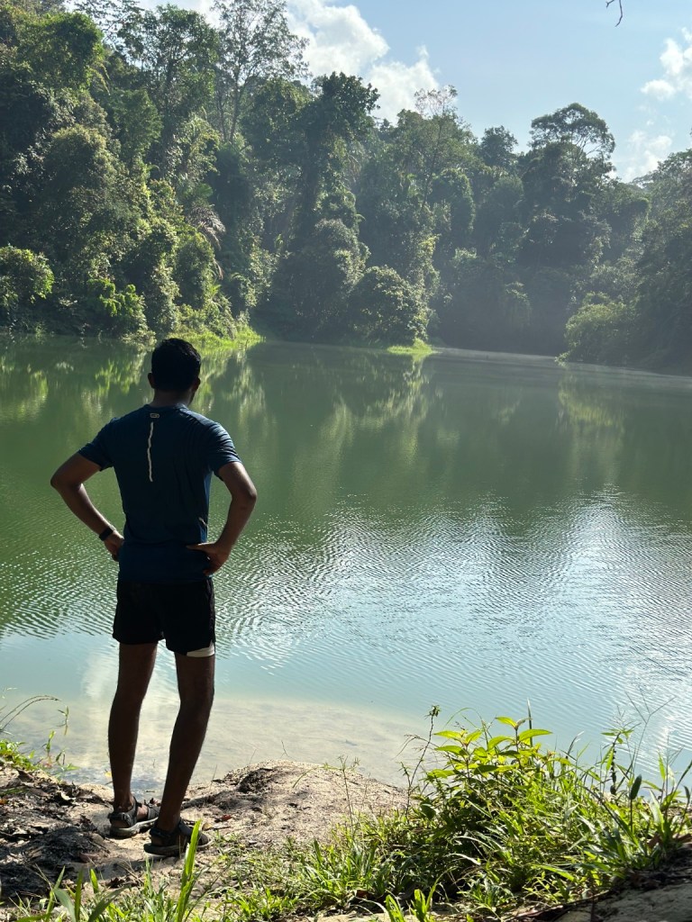 Thana standing beside calm water facing the surrounding forest