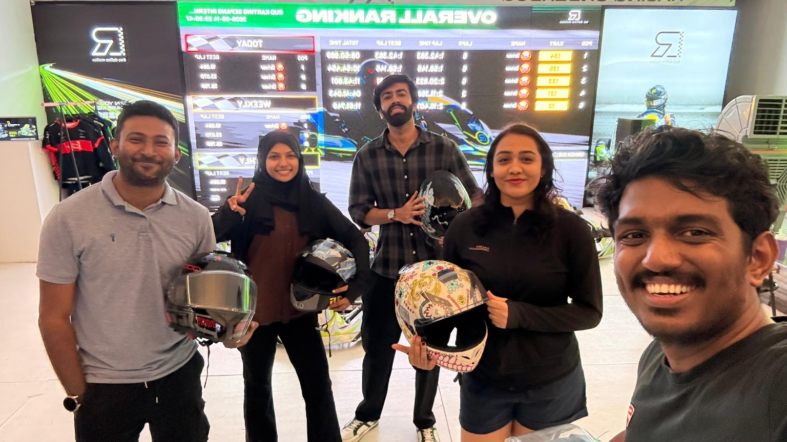Thana and colleagues holding helmets beside the karting timing screen