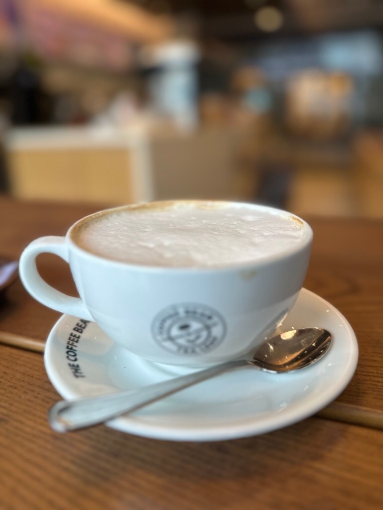 A white Coffee Bean cup of frothy coffee with a spoon on its saucer