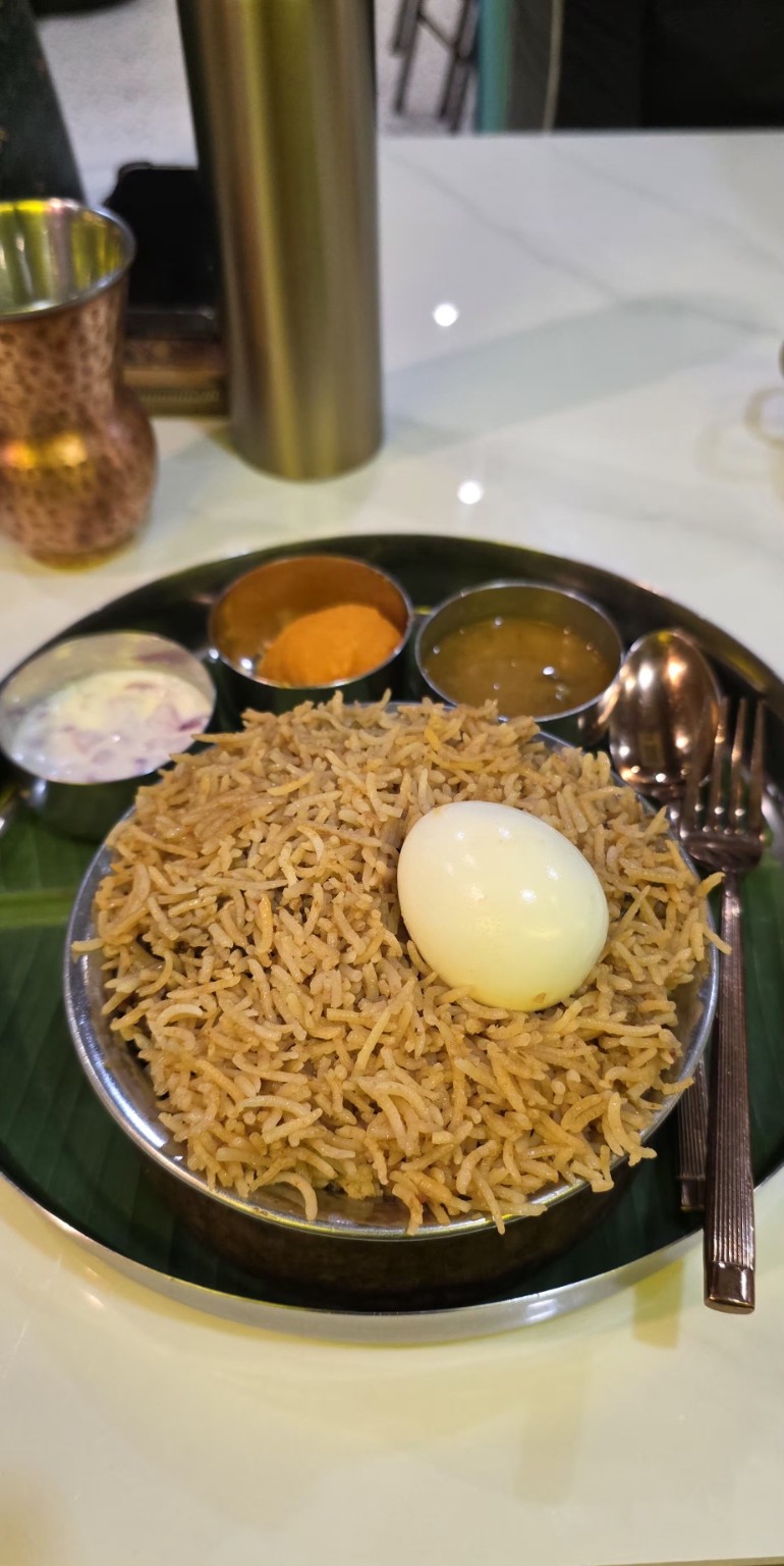 Briyani served in a metal bowl with a boiled egg and small dishes of accompaniments.